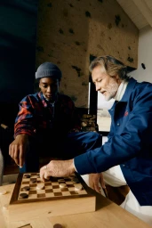 Two men playing checkers indoors, one wearing a blue beanie and plaid shirt, the other an older man in a blue jacket.