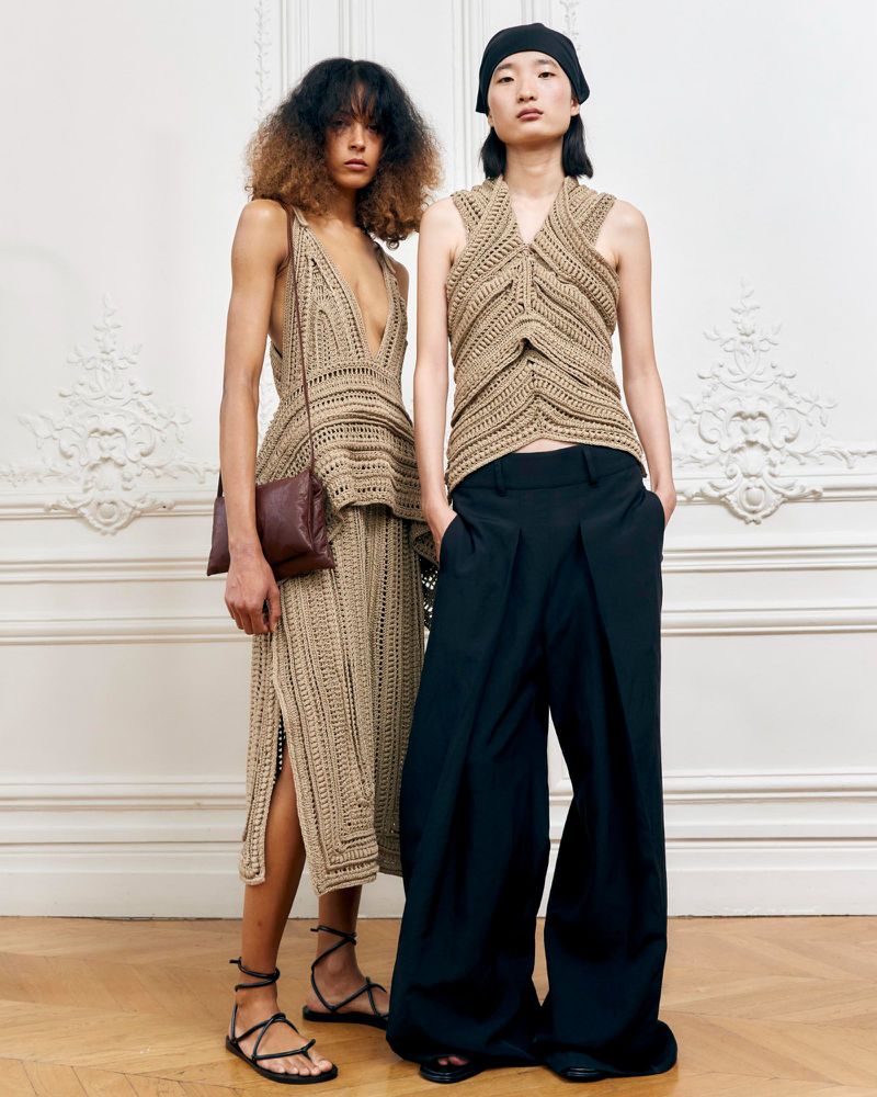Two fashion models pose in knitwear outfits in a studio against a white wall, one in a tan top and skirt, the other in black pants and a tan top.