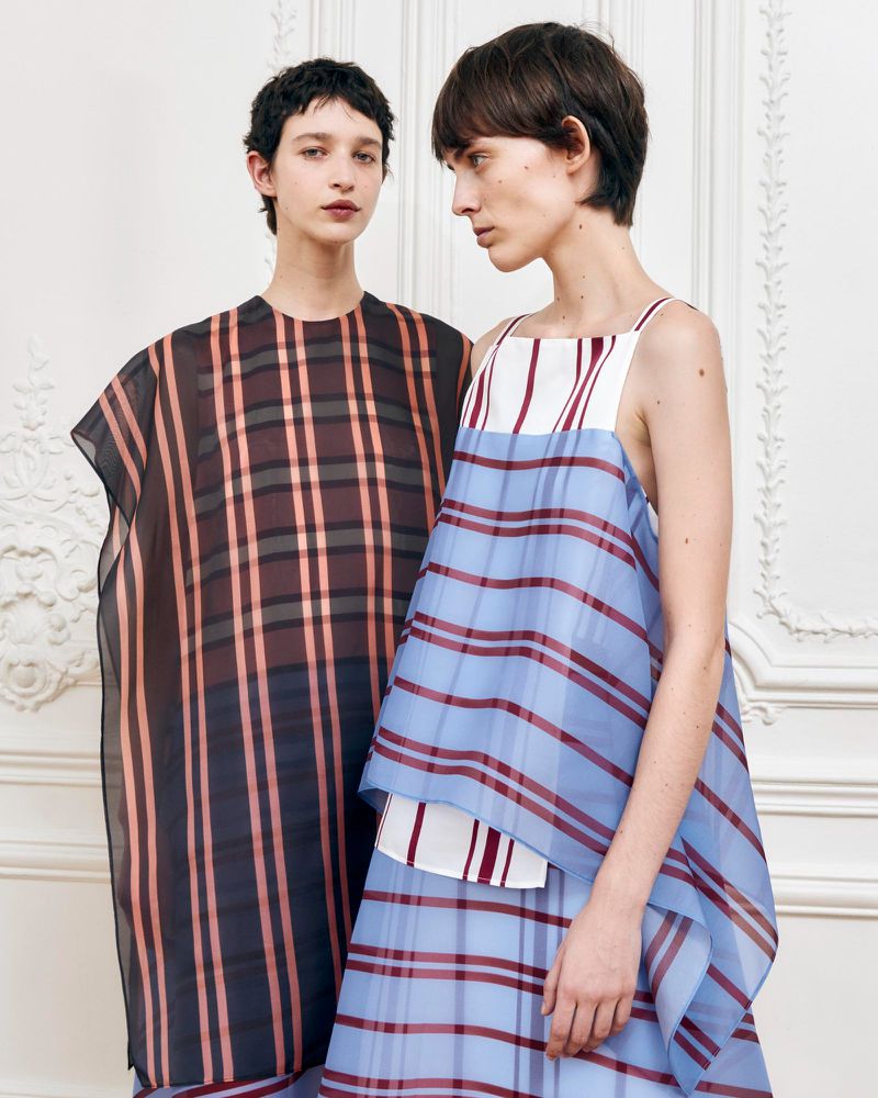 Two women pose indoors wearing sheer, plaid tops in blue and brown tones, standing against a decorative, patterned white wall.