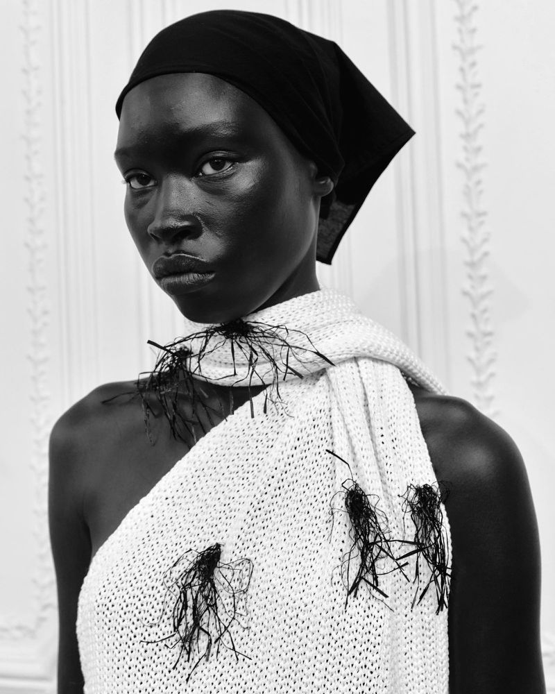 Portrait of an adult Black woman wearing a black headscarf and an off-the-shoulder knitted white garment with decorative black tassels.