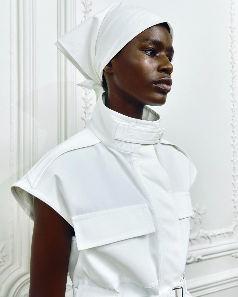 Elegant portrait of a Black woman with a white headscarf and a white, sleeveless safari top, standing indoors against a textured white wall.