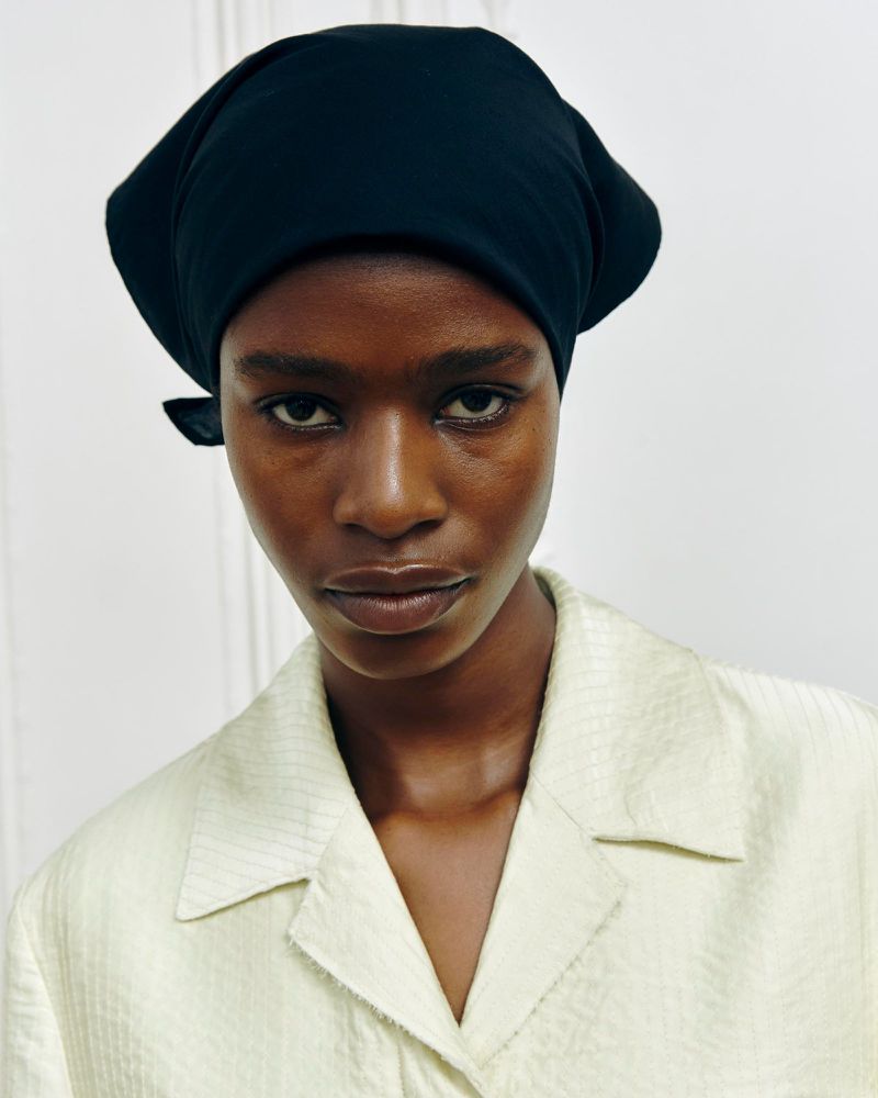 Close-up portrait of an adult woman with dark skin wearing a black headscarf and a light green button-down shirt against a white background.