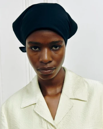 Close-up portrait of an adult woman with dark skin wearing a black headscarf and a light green button-down shirt against a white background.