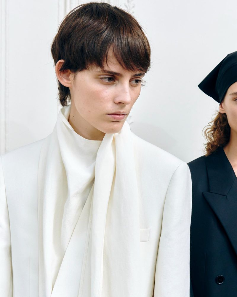 Close-up of a young adult female model with short brown hair and a white jacket with a scarf, standing next to a model in black.