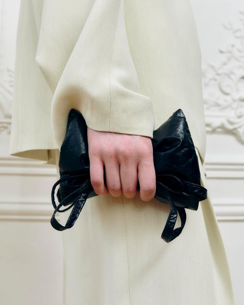 Person in light-colored suit holds black leather clutch with gathered details against a pale ornamental background.
