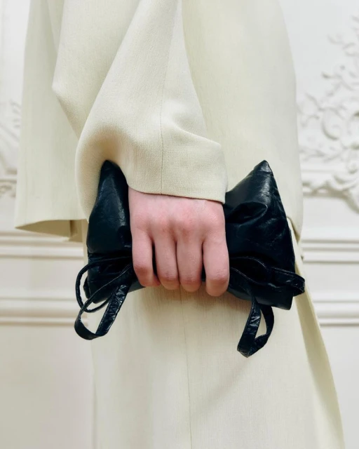 Person in light-colored suit holds black leather clutch with gathered details against a pale ornamental background.