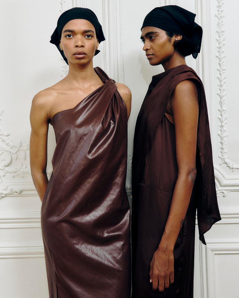 Two fashion models in brown dresses and black headscarves pose indoors against a decorative white wall, showcasing modern apparel.