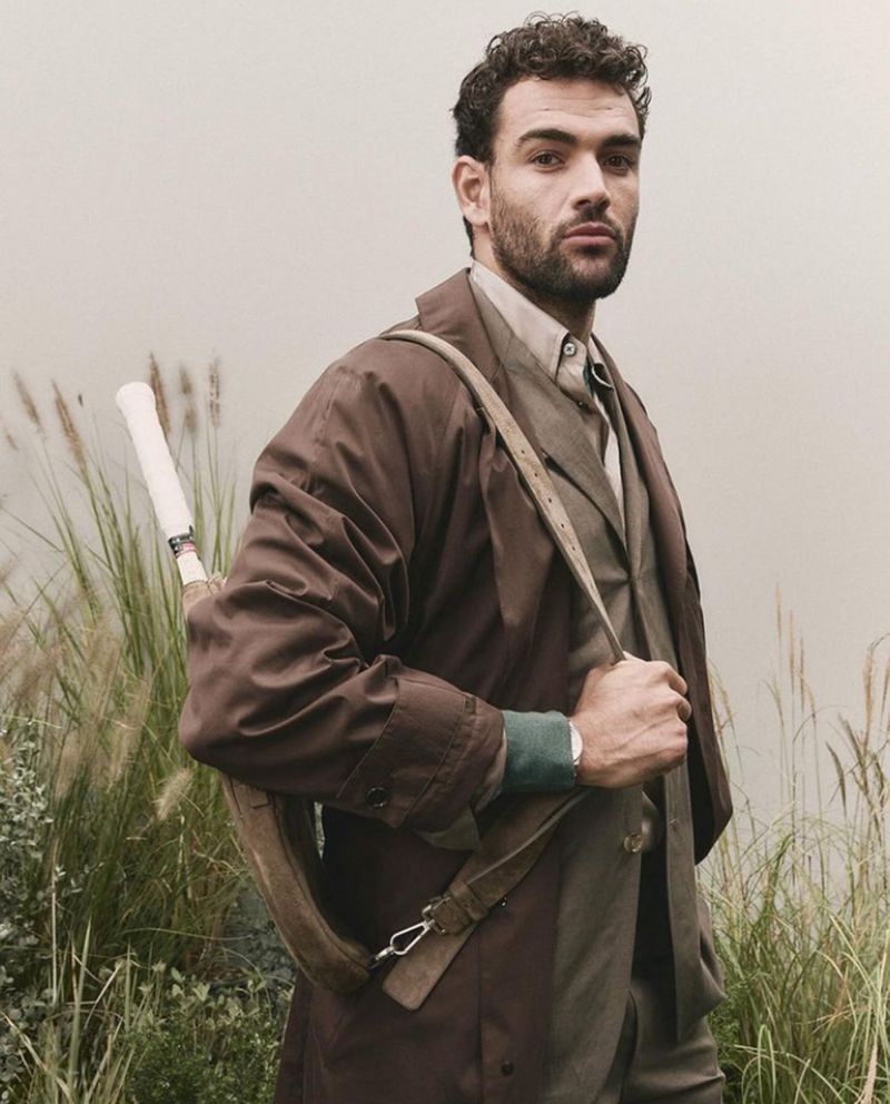 Adult male outdoors in olive suit, overcoat, holding tennis racket, posing near tall reeds and grass in soft daylight.