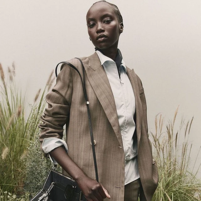 A Black female model in a brown double-breasted suit and black purse poses in a green field with a neutral wall backdrop.