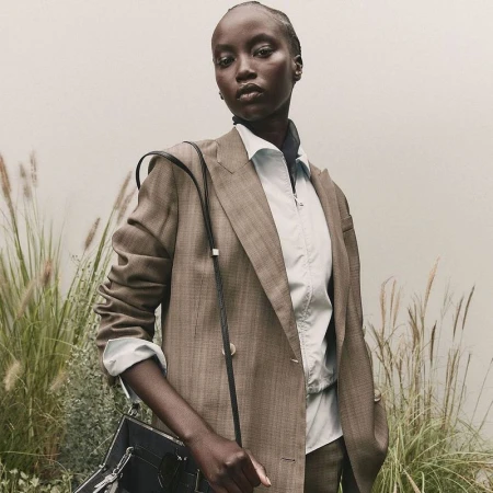 A Black female model in a brown double-breasted suit and black purse poses in a green field with a neutral wall backdrop.