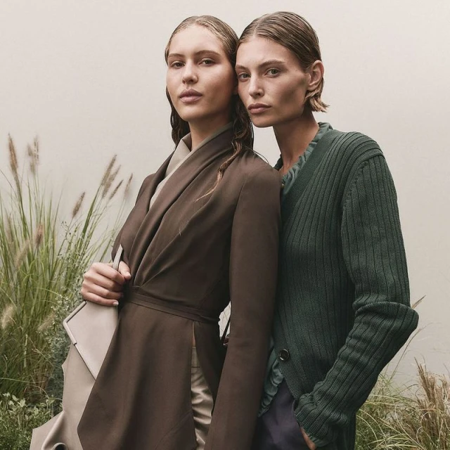 Two women in fashionable earth-tone outerwear pose in a medium shot against a neutral backdrop with natural grasses.