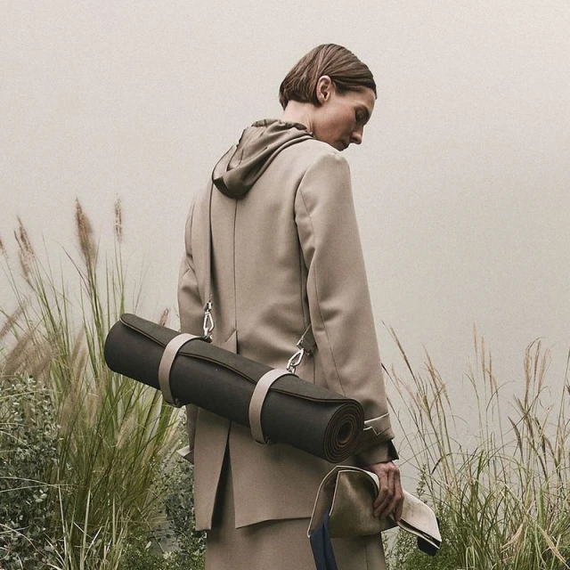 Adult female model carrying a rolled mat and a bag, wearing a beige jacket with a hood in an outdoor grassy environment.