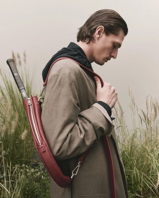 Adult male in coat carries a burgundy tennis racket bag in grassy outdoor setting, showcasing fashion and sport lifestyle.