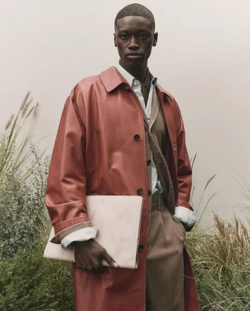 Black man wearing a red coat holding a light-colored clutch in front of grassy foliage for BOSS S/S 25.