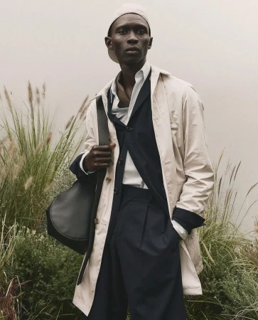 Black male model wearing light coat, dark suit, cap, and holding black leather shoulder bag amidst tall grass.