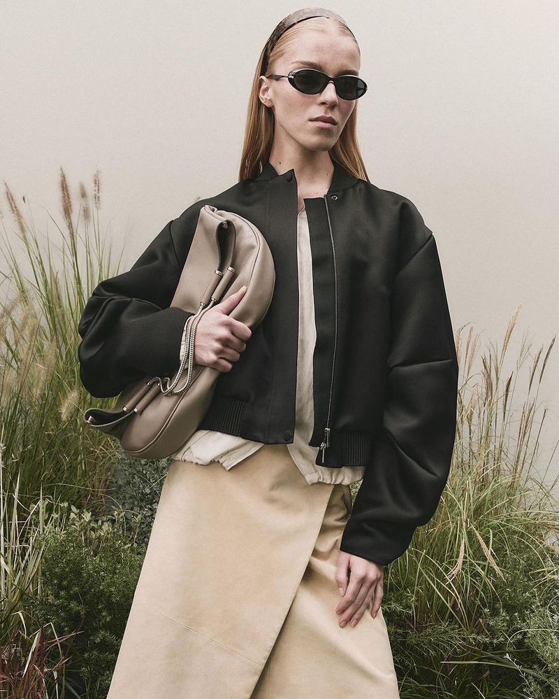 Woman in black jacket and beige skirt posing with large beige clutch in outdoor setting with dry grass and muted light.