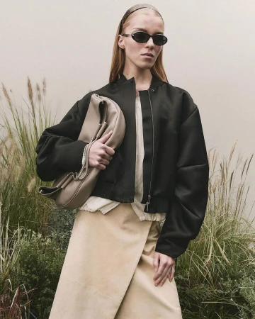 Woman in black jacket and beige skirt posing with large beige clutch in outdoor setting with dry grass and muted light.