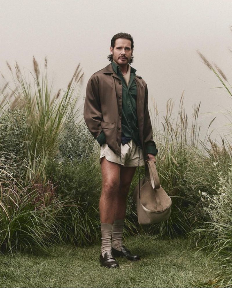 Man standing outdoors among tall grass wearing a brown jacket, green shirt, shorts, and carrying a bag for BOSS S/S 25.