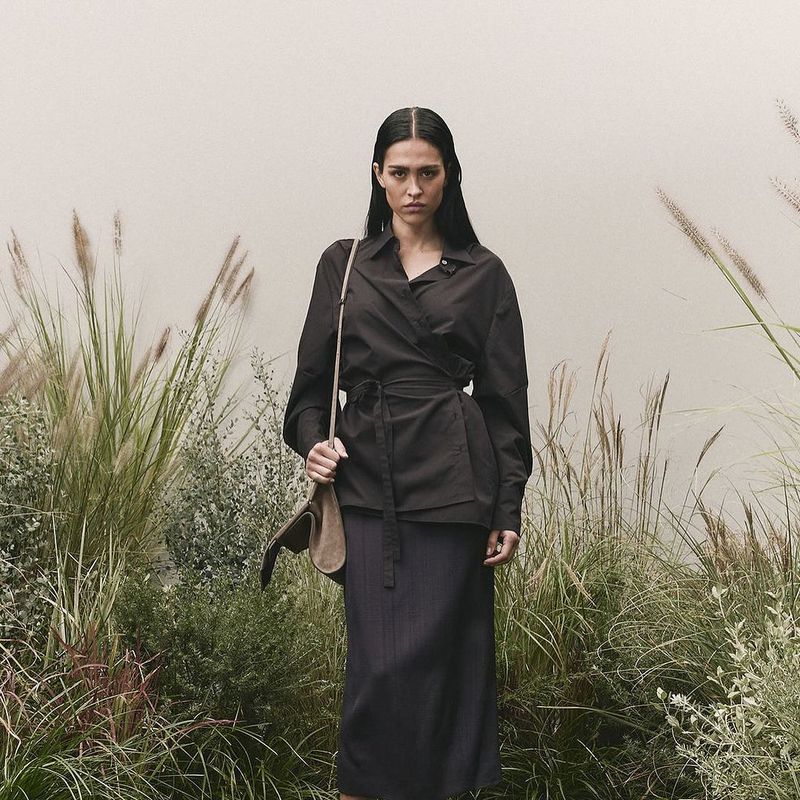 Adult woman in a dark wrap shirt and skirt with a shoulder bag, standing outdoors surrounded by tall grass and foliage against a light wall.