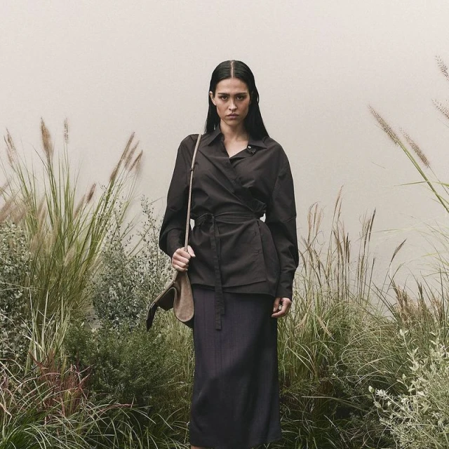 Adult woman in a dark wrap shirt and skirt with a shoulder bag, standing outdoors surrounded by tall grass and foliage against a light wall.