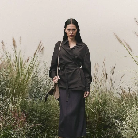 Adult woman in a dark wrap shirt and skirt with a shoulder bag, standing outdoors surrounded by tall grass and foliage against a light wall.