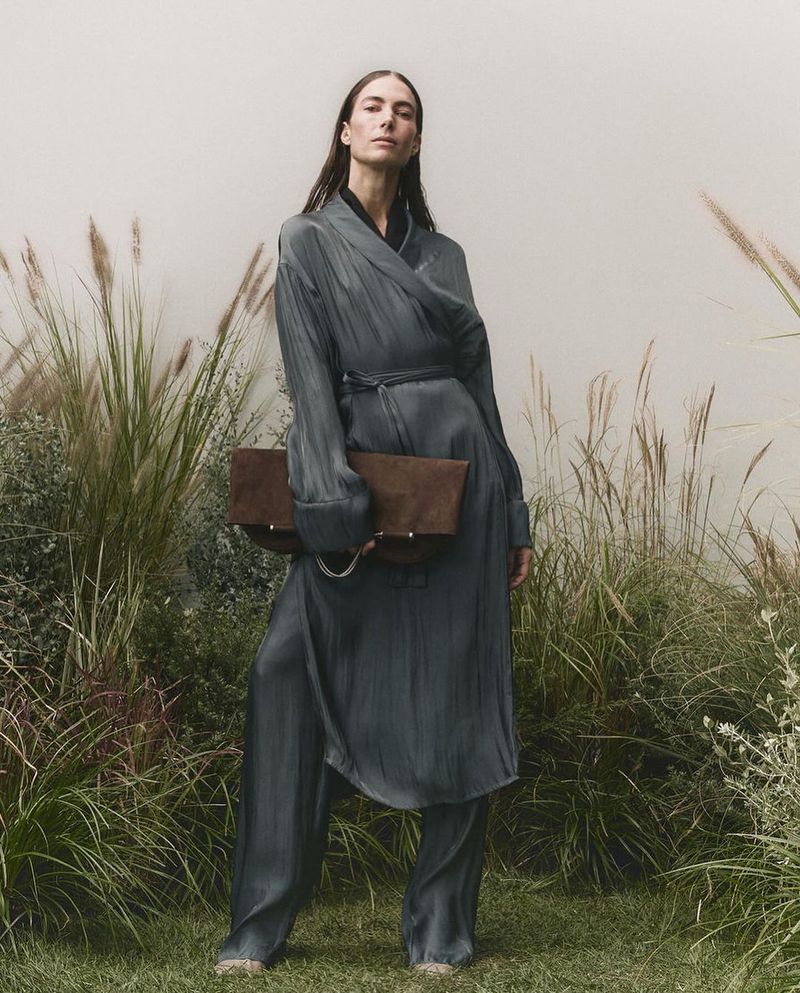 Elegant woman in flowing blue-grey robe and pants, holding a brown clutch, poses outdoors amid tall grasses and soft natural light.