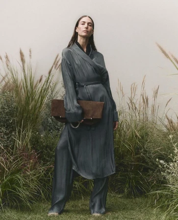 Model in a grey satin wrap coat holding a brown clutch standing among tall grass in a campaign for BOSS S/S 25.