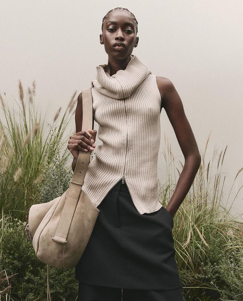 Fashionable adult African woman poses with a large beige shoulder bag in a sleeveless knit top and black skirt outdoors.