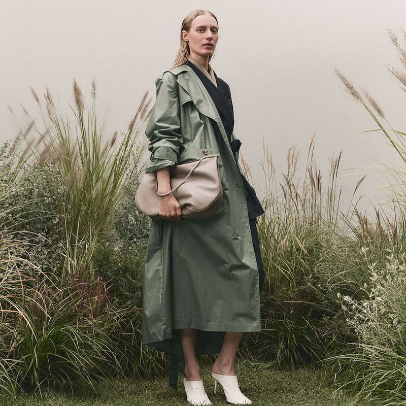 A woman with blonde hair and a green trench coat is holding a handbag while wearing heels outdoors amongst tall grassy reeds.