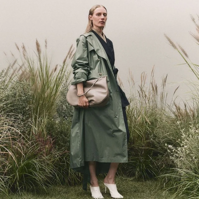 A woman with blonde hair and a green trench coat is holding a handbag while wearing heels outdoors amongst tall grassy reeds.