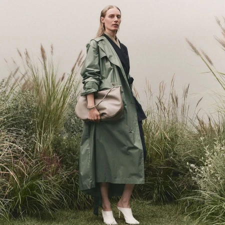 A woman with blonde hair and a green trench coat is holding a handbag while wearing heels outdoors amongst tall grassy reeds.