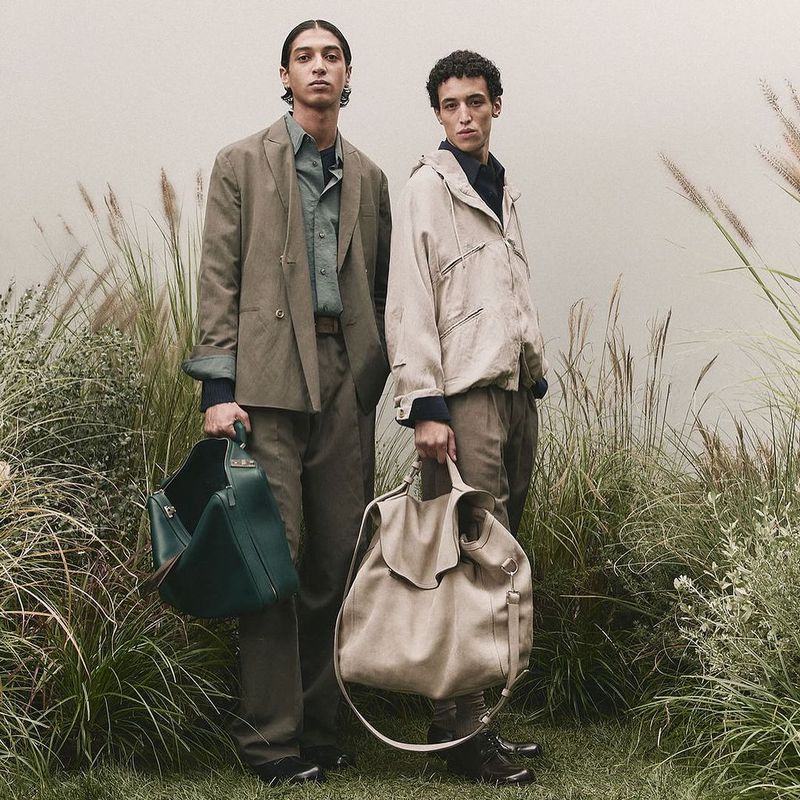 Two young adult male models in muted-tone casual fashion hold large handbags amidst a field of tall grass, shot in soft, diffused lighting.