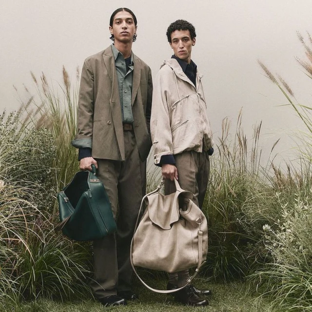Two young adult male models in muted-tone casual fashion hold large handbags amidst a field of tall grass, shot in soft, diffused lighting.
