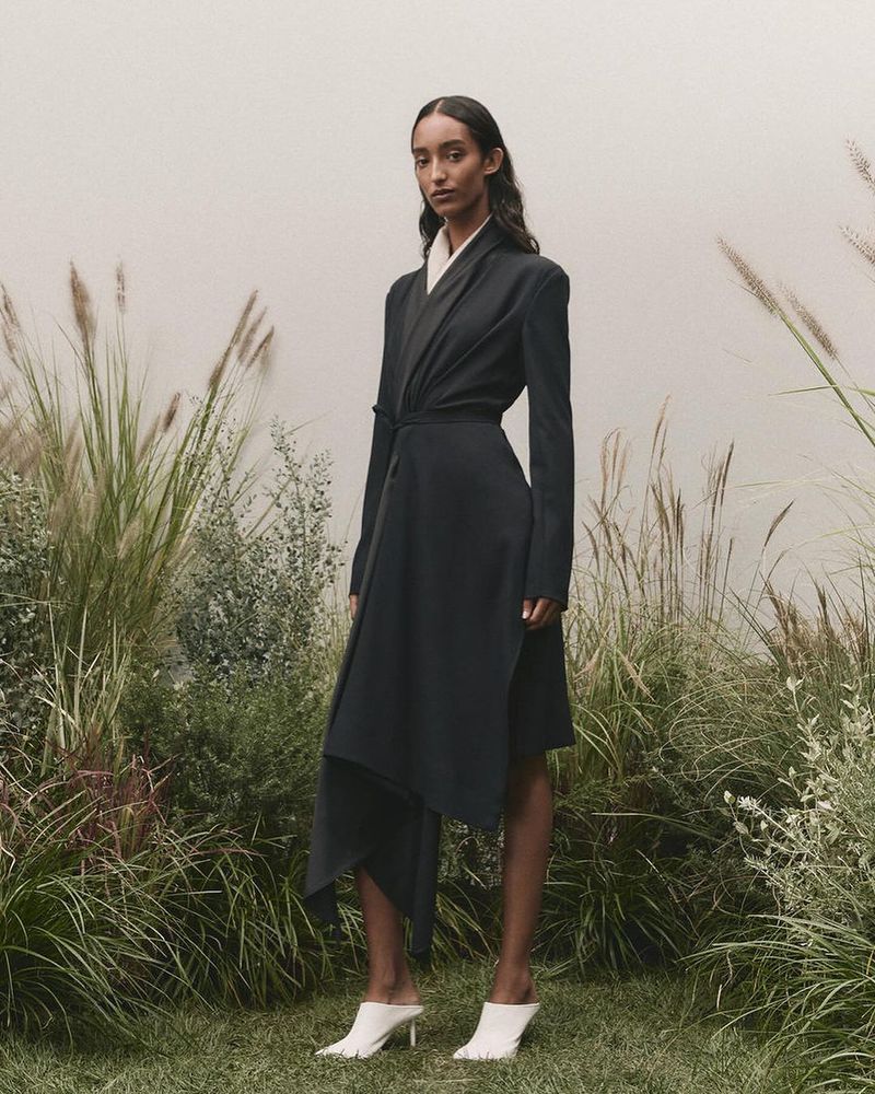Fashion model poses in a long-sleeve black dress with white heeled mules in a grassy field. Tall beige wild grasses surround the model.