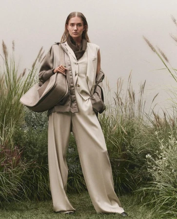 Model standing in tall grass wearing a tailored beige suit vest, wide-leg trousers, and carrying a large shoulder bag.