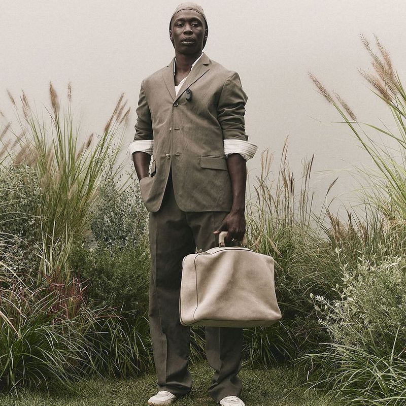 Full-length shot of a Black man in a suit, head covering, and sneakers, holding a duffel bag, standing in a field of tall grass.