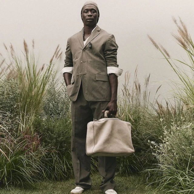 Full-length shot of a Black man in a suit, head covering, and sneakers, holding a duffel bag, standing in a field of tall grass.