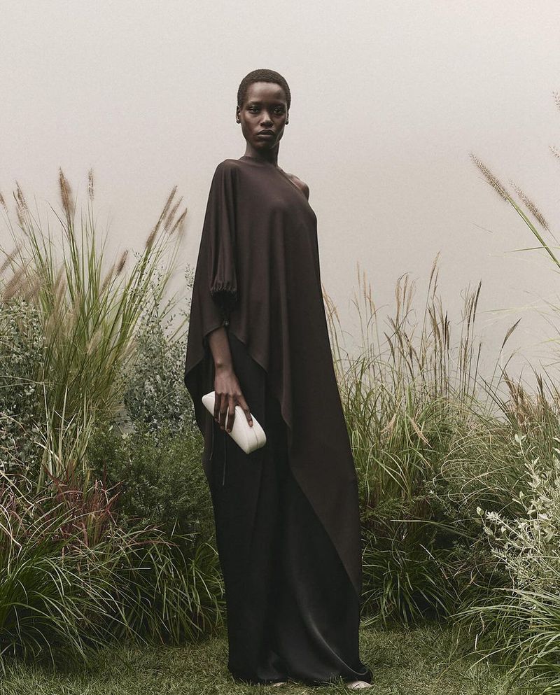 An African woman in a brown dress holds a white clutch, posing outdoors among tall grasses in a fashion editorial shot on a cloudy day.