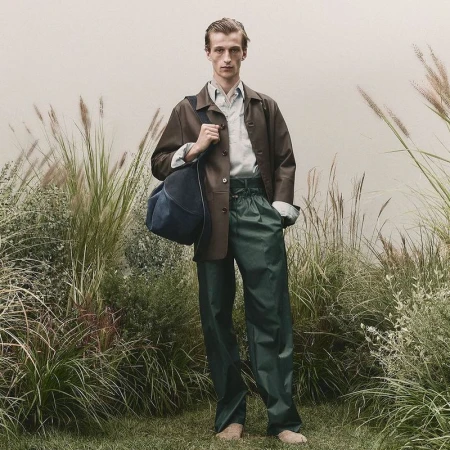 A fair-skinned male model poses in a grassy field, wearing a brown jacket, green pants, and carrying a blue duffel bag on his shoulder.