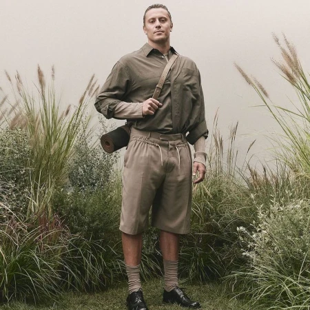 A blond male in an olive shirt and khaki shorts poses outdoors amidst tall grass and a neutral backdrop, showcasing a casual yet put-together look.