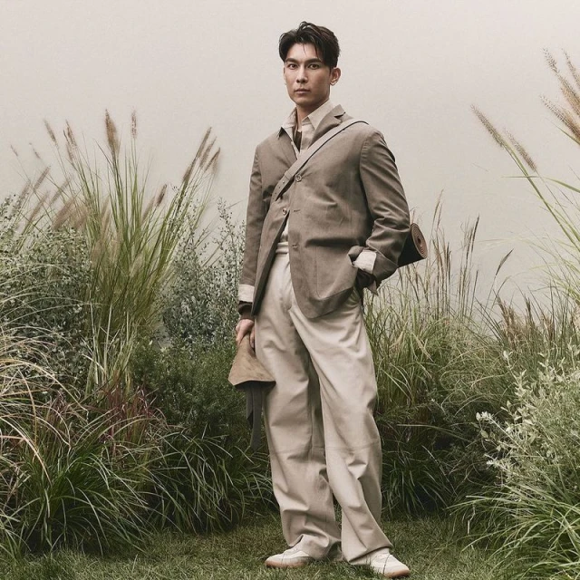 Man in suit standing in grassy field wearing shoulder bag and holding hat, looking forward. Tall grass and neutral wall background.