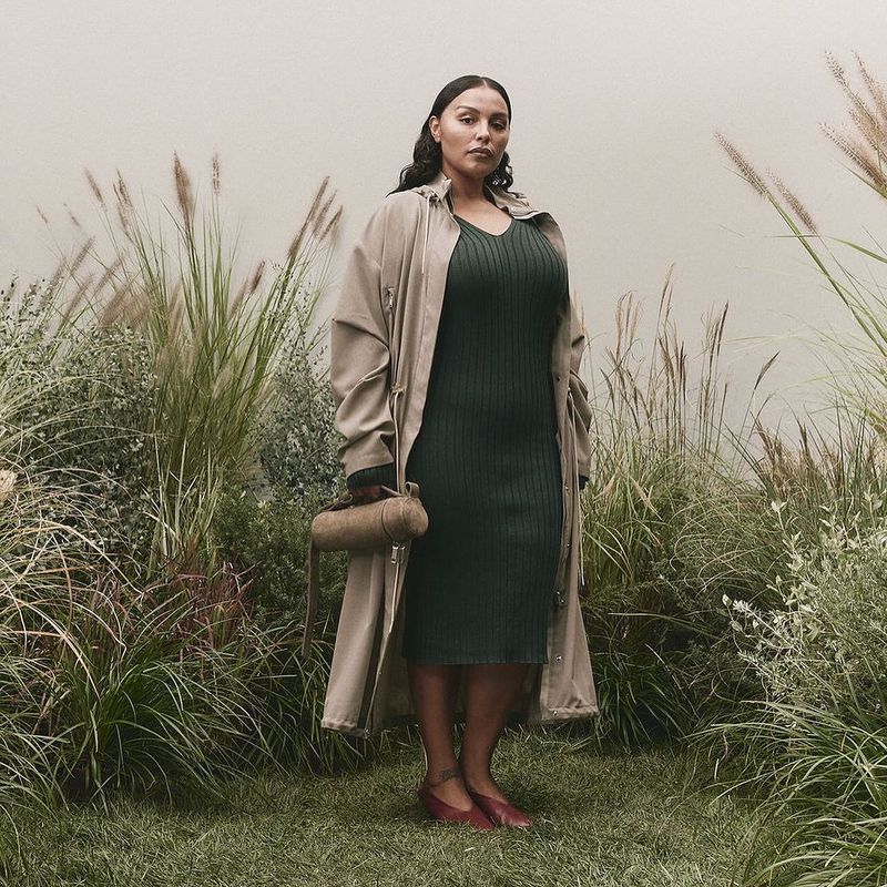 Adult female stands in front of a wall surrounded by tall grass wearing a dark green dress, tan trench coat, and holding a cylinder purse.
