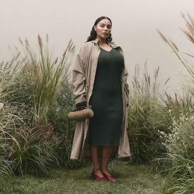 Adult female stands in front of a wall surrounded by tall grass wearing a dark green dress, tan trench coat, and holding a cylinder purse.
