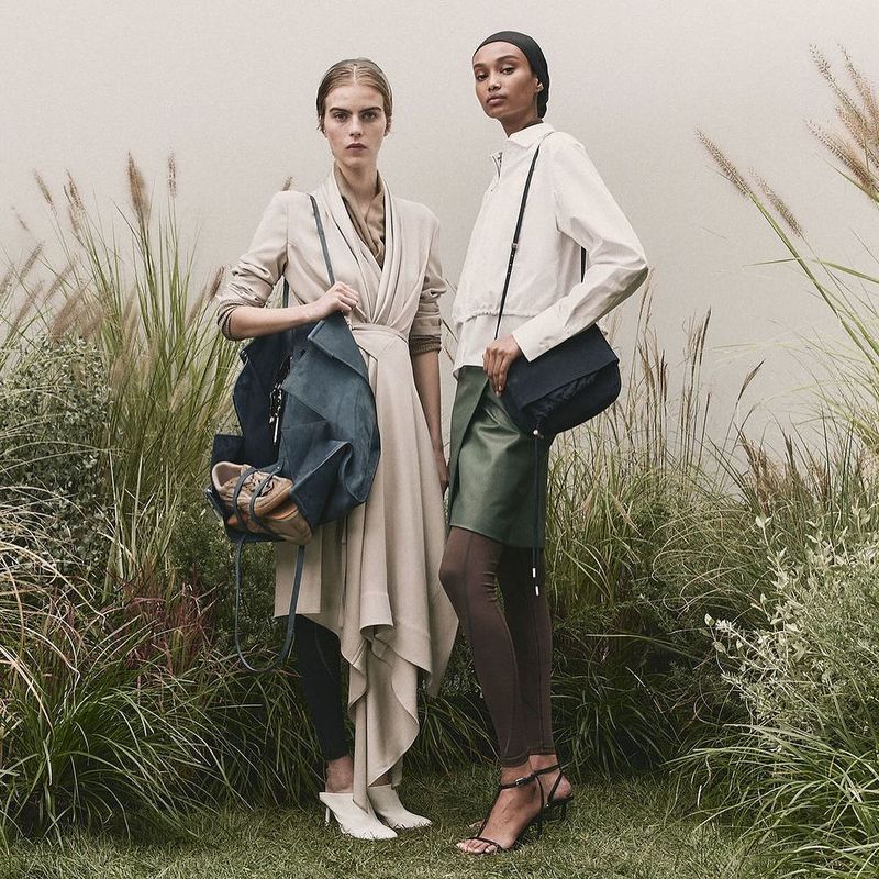 Two fashion models pose in outdoor setting, one with beige dress and backpack, the other in white shirt, green skirt, and shoulder bag.