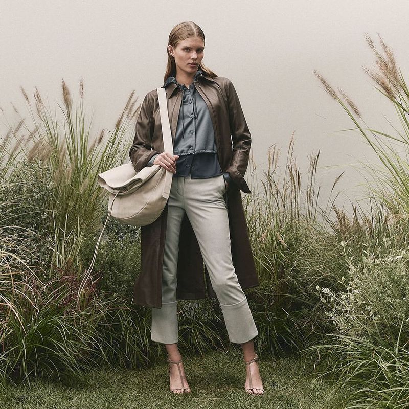 Fashionable woman posing in a stylish brown leather trench coat, blue blouse, light pants, and beige bag in a lush outdoor setting.