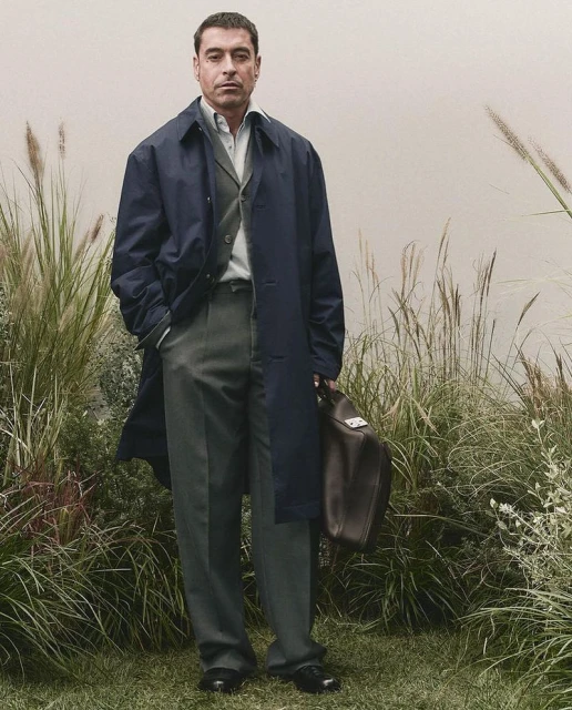 Full-length shot of an adult male wearing a navy overcoat, dress shirt, and grey slacks, holding a leather duffel bag in a grassy outdoor setting.