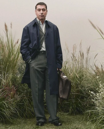 Full-length shot of an adult male wearing a navy overcoat, dress shirt, and grey slacks, holding a leather duffel bag in a grassy outdoor setting.