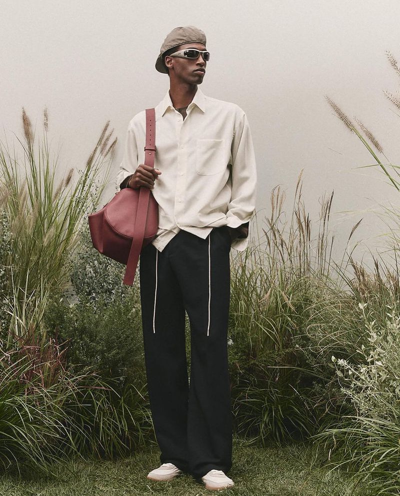 Stylish man in shirt, trousers, beret, sunglasses, and handbag poses outdoors against muted backdrop with ornamental grasses.