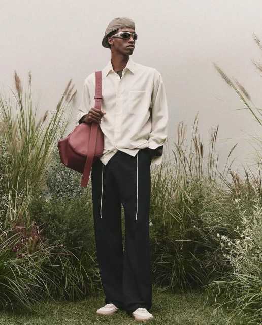Stylish man in shirt, trousers, beret, sunglasses, and handbag poses outdoors against muted backdrop with ornamental grasses.