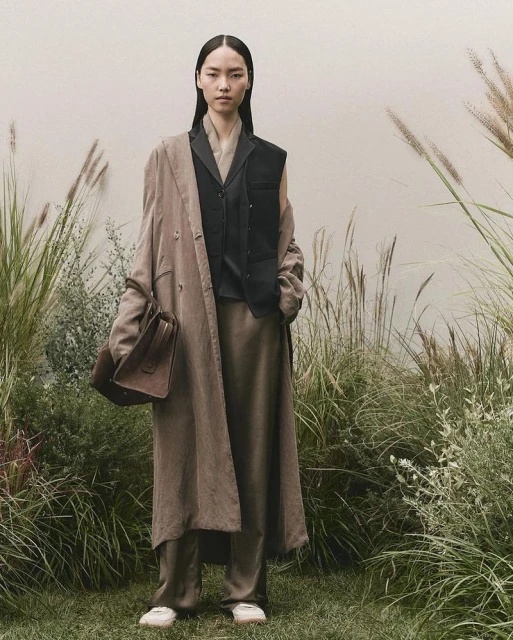 Elegant Asian woman in flowing brown coat, black vest, and neutral trousers stands in a grassy field with tall wheat-like plants.